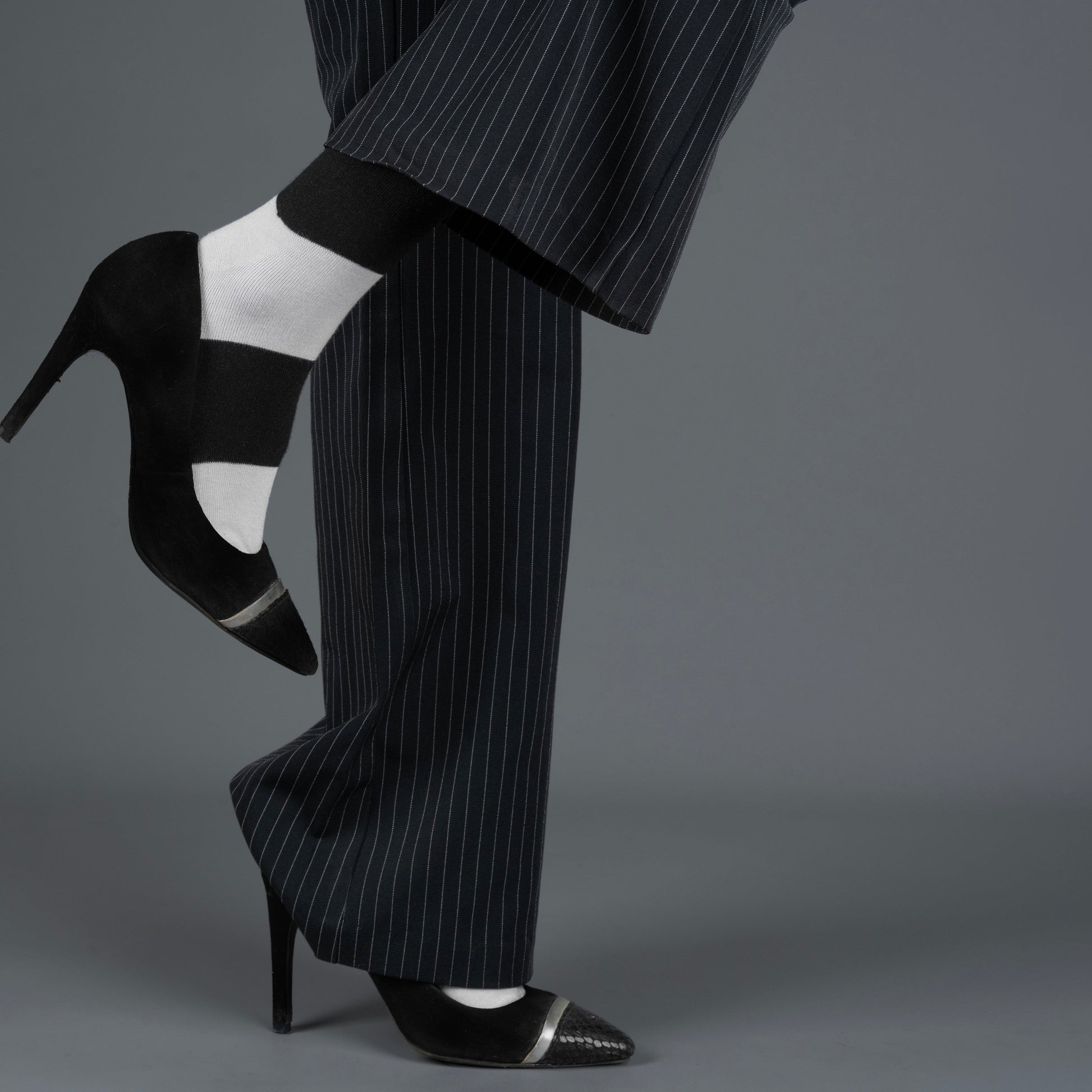 Black high-heeled shoes with white soles worn with a black pinstripe suit and black and white bamboo socks on a gray background