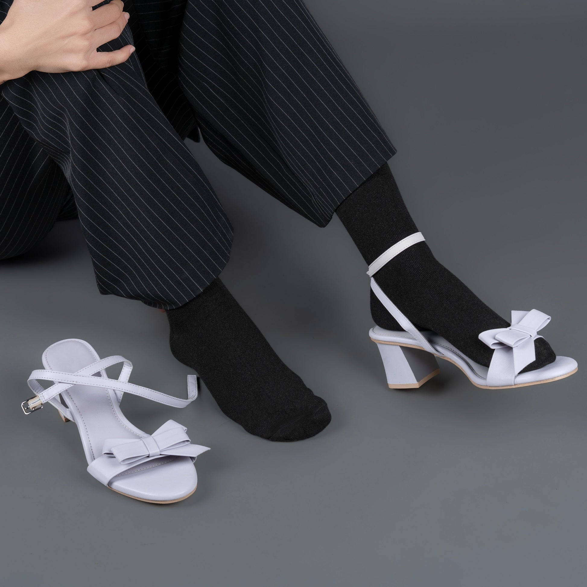 Person wearing black pants, black bamboo socks and white sandals with bows on a gray background