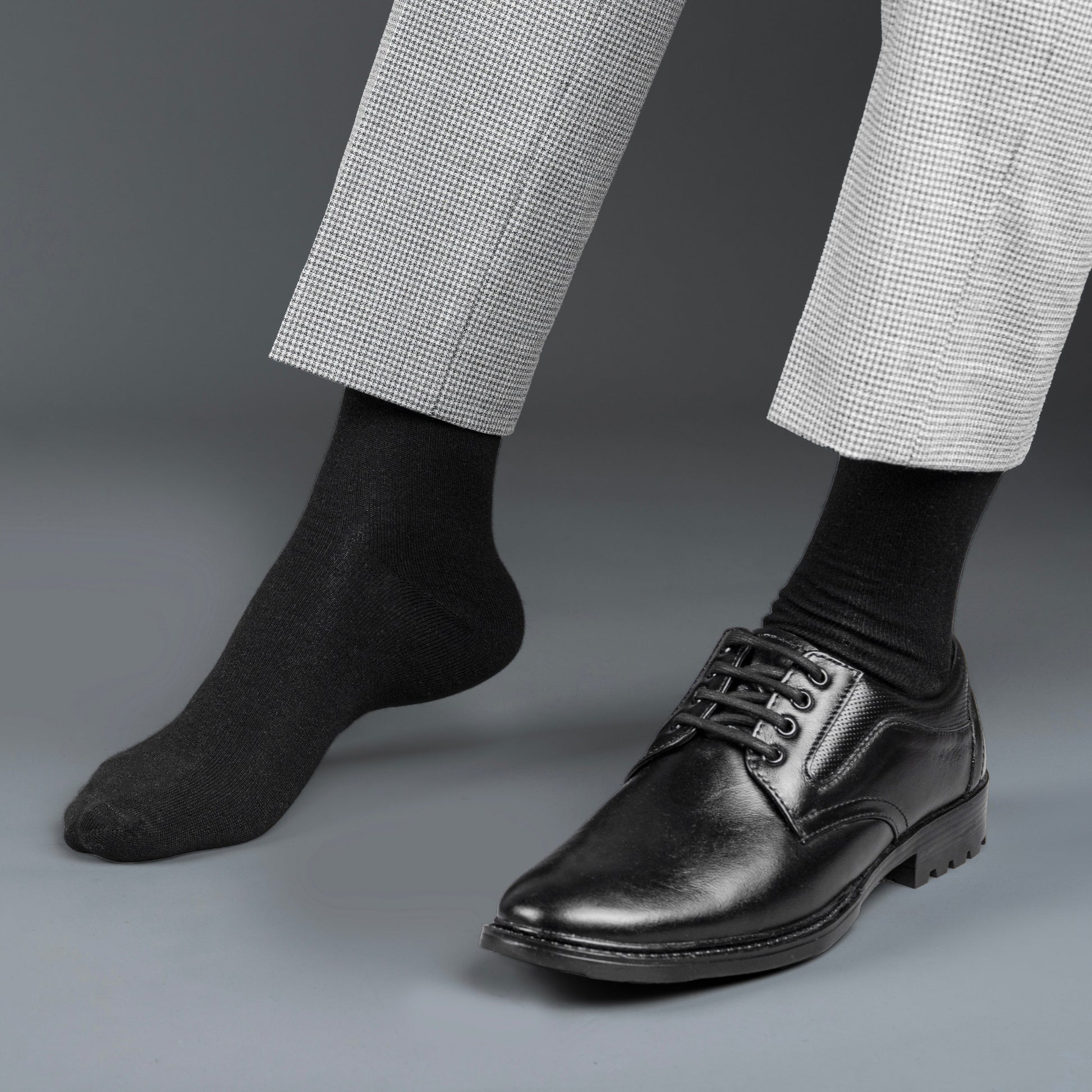 Black dress shoes with white laces worn with gray checkered pants and black bamboo socks on a gray background